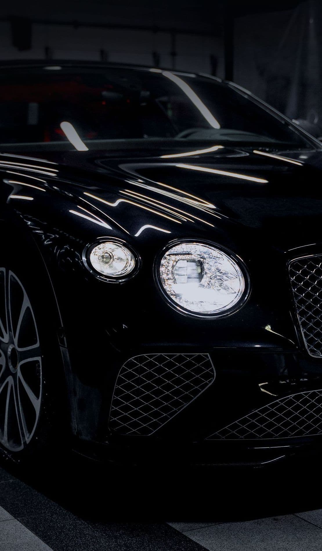 A black Bentley Continental GT in a dark preparation bay, headlamps on.