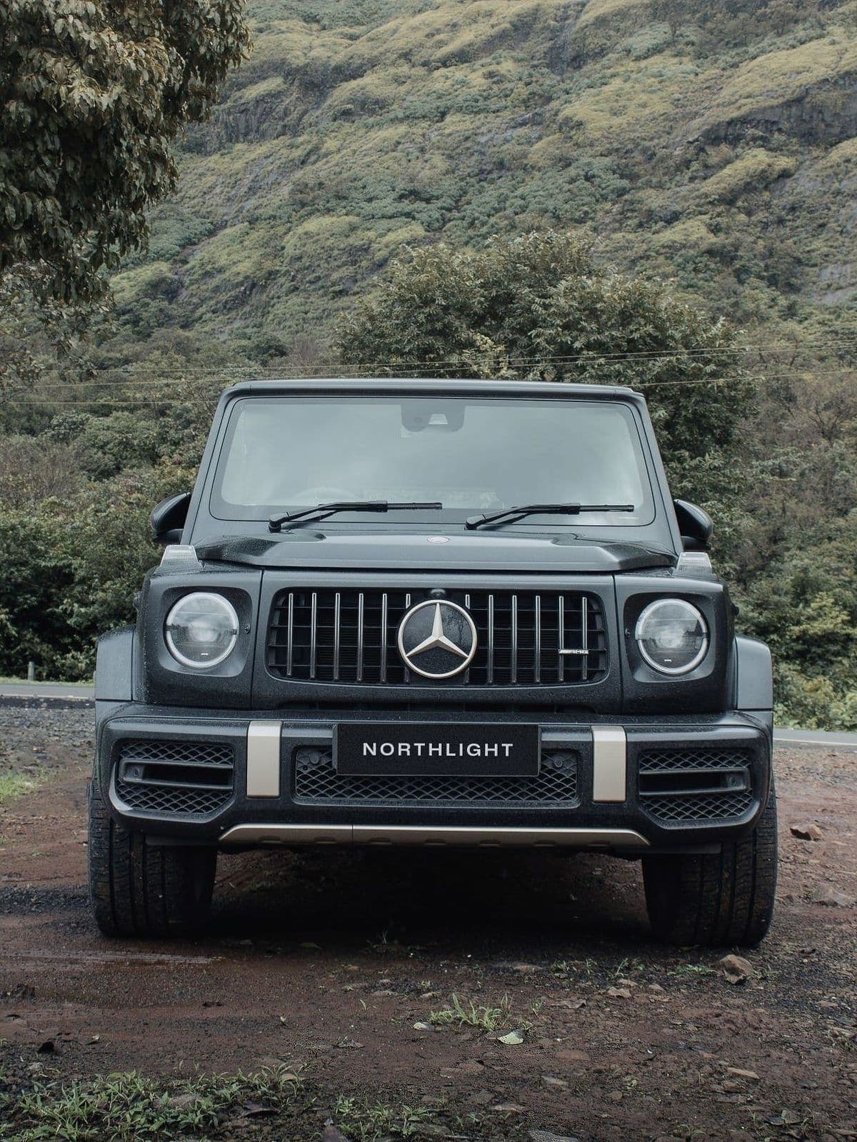 A matte black-grey G 63 on a red-earth track below green hills.