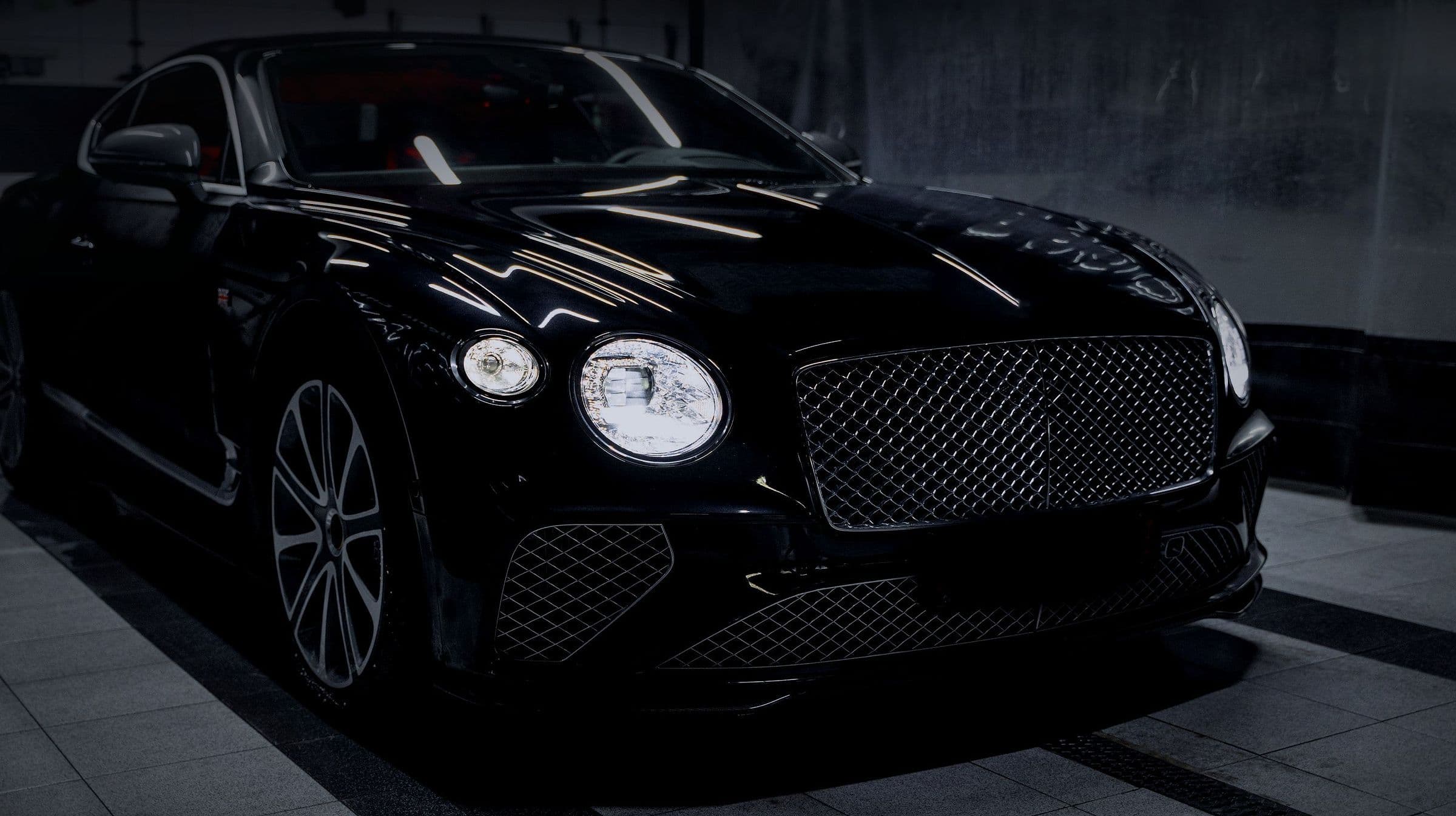 A black Bentley Continental GT in a dark preparation bay, headlamps on.