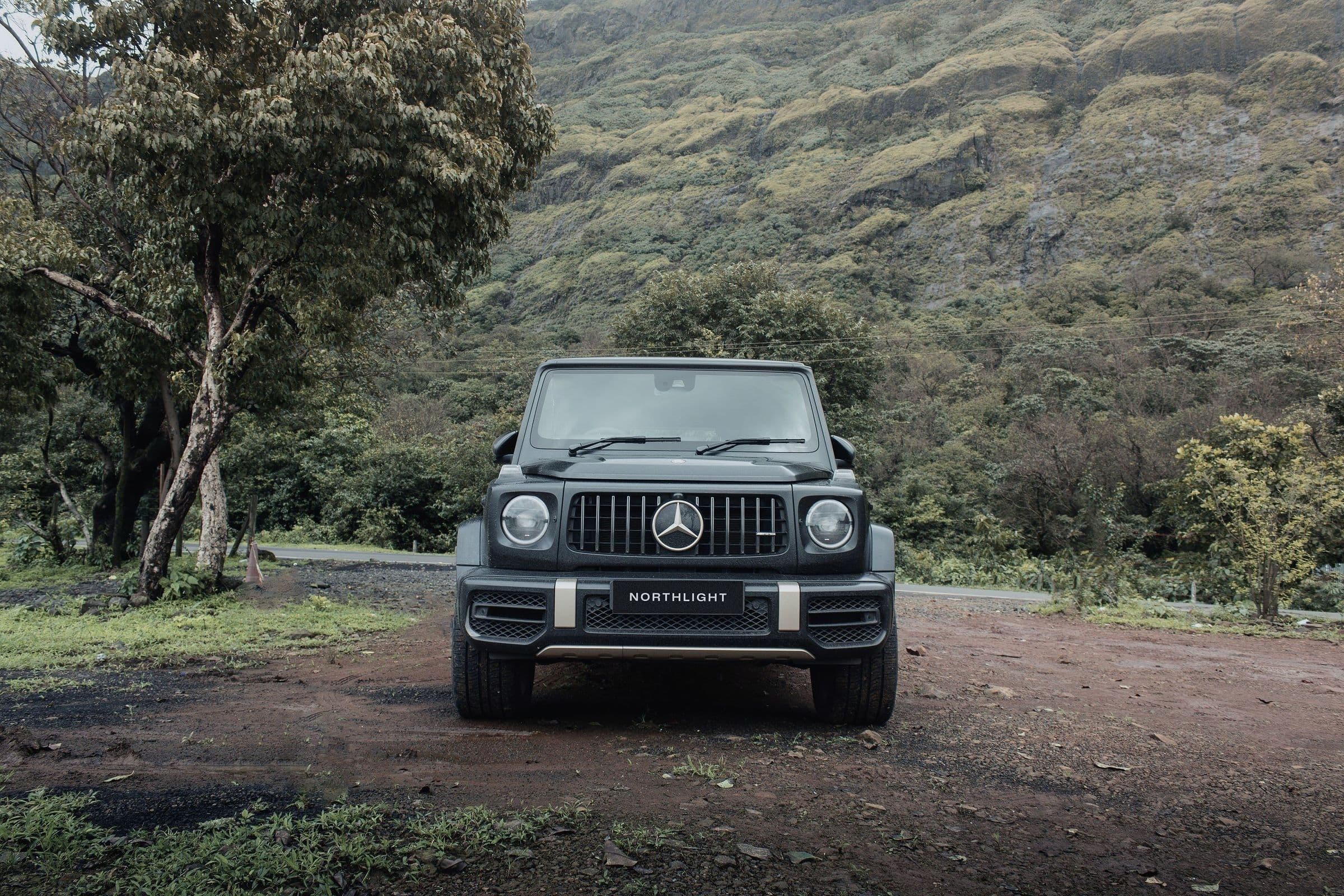 A matte black-grey G 63 on a red-earth track below green hills.