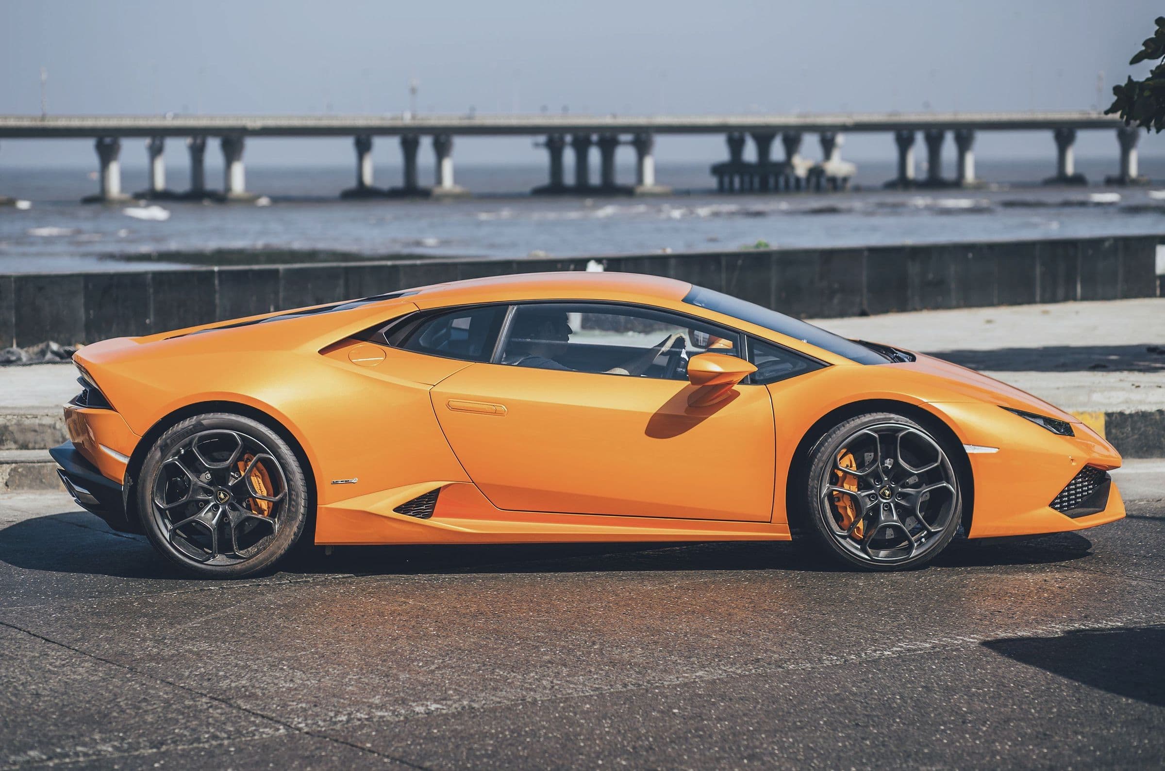 An orange Lamborghini Huracán in profile on a seafront road, a sea bridge behind.