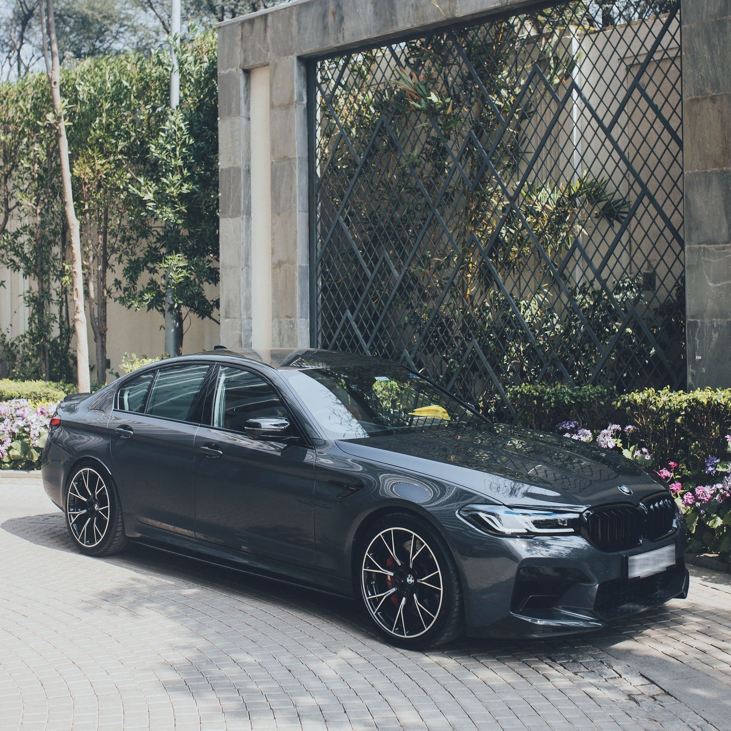 A dark grey BMW M5 in a stone-walled driveway with flowering beds.
