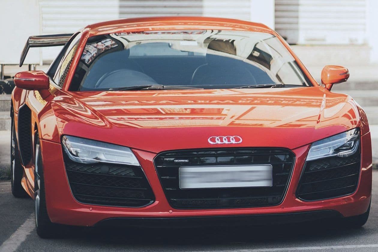 A red Audi R8 with a fixed rear wing, parked in front of closed shutters.